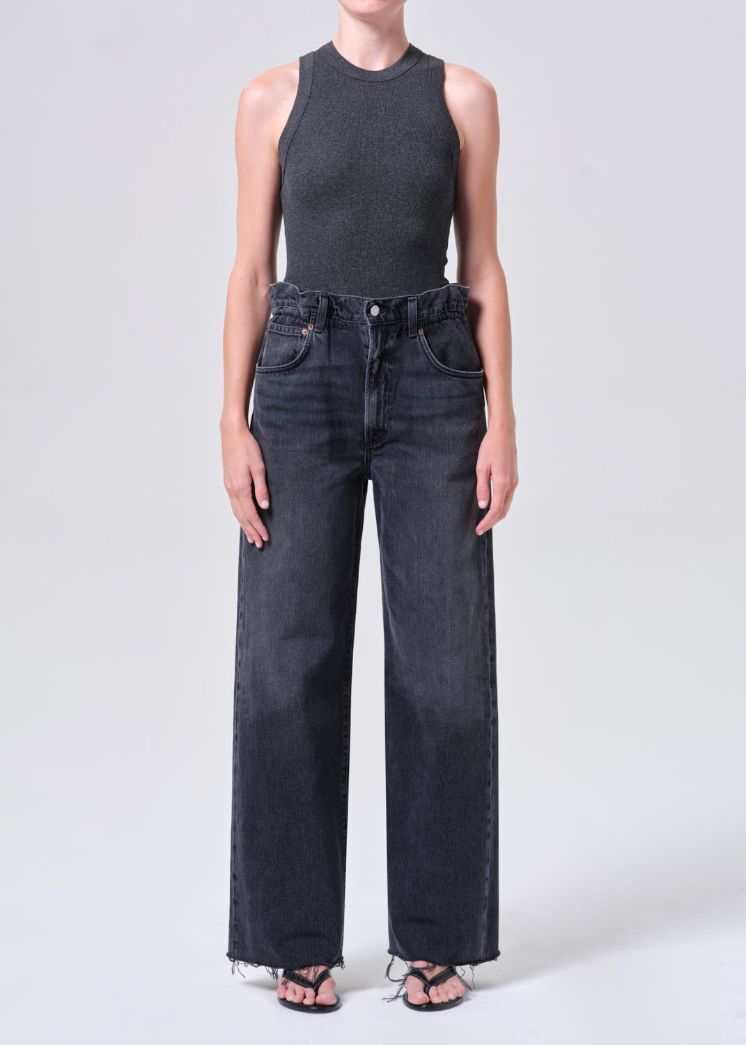 Person wearing a dark gray sleeveless top and black high-waisted jeans on a light gray background