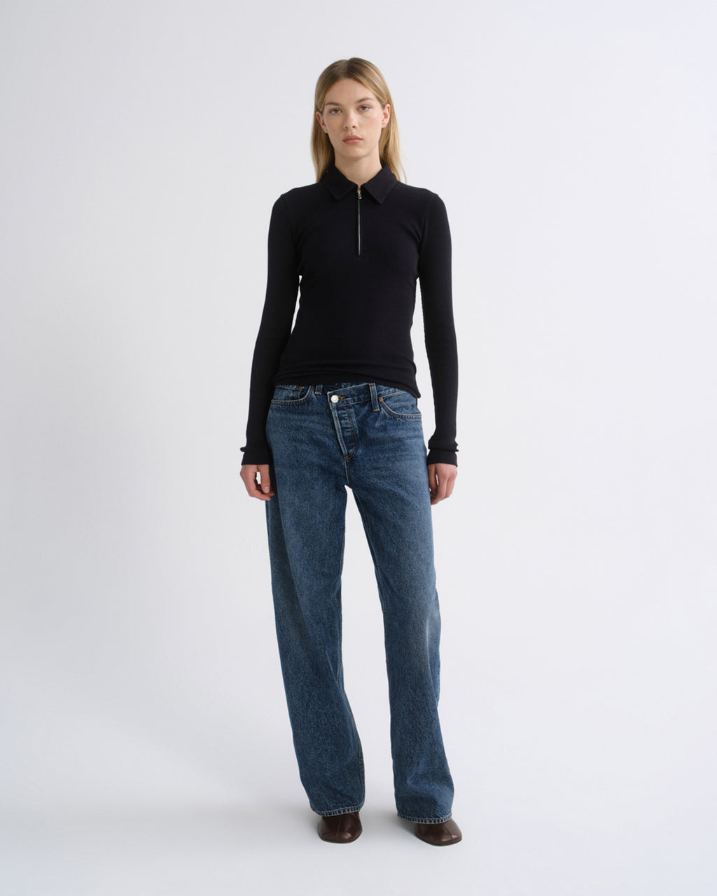 A model stands against a light gray studio background wearing a black long-sleeve zip-neck top and blue wide-leg jeans.