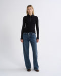 A model stands against a light gray studio background wearing a black long-sleeve zip-neck top and blue wide-leg jeans.