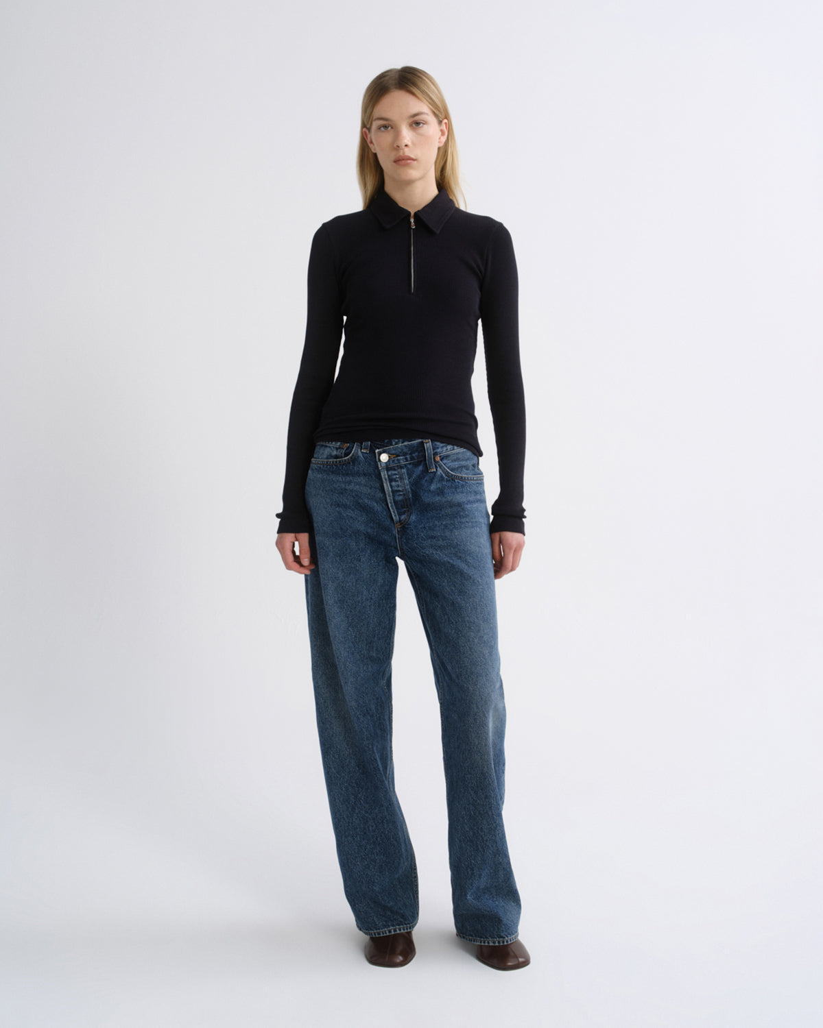 A model stands against a light gray studio background wearing a black long-sleeve zip-neck top and blue wide-leg jeans.