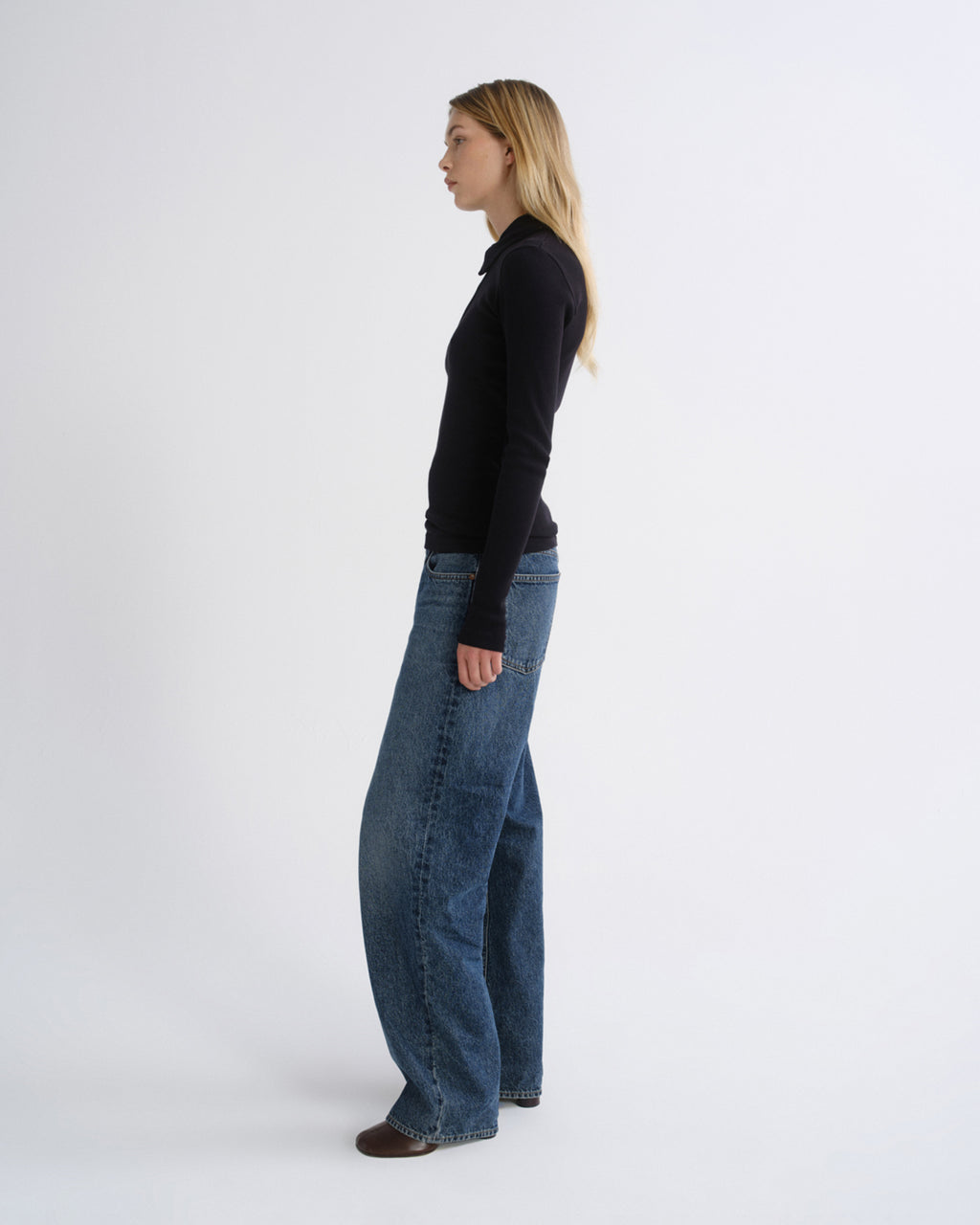 A woman standing in profile wearing a black long-sleeve top and wide-leg blue jeans against a plain light gray studio background.