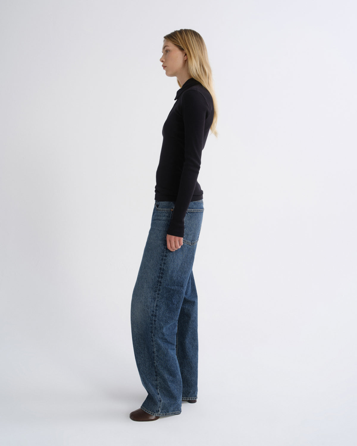A woman standing in profile wearing a black long-sleeve top and wide-leg blue jeans against a plain light gray studio background.