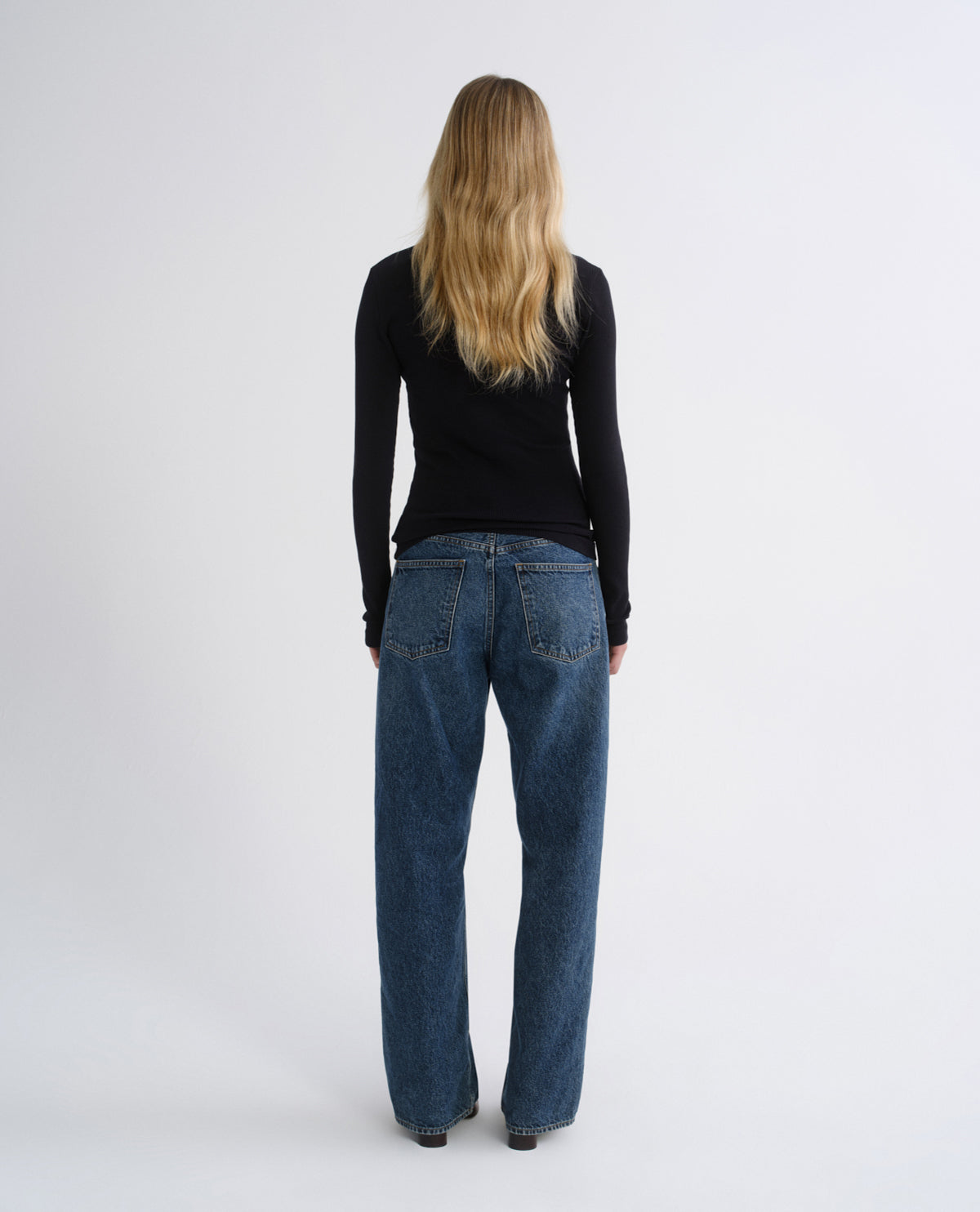 A woman viewed from behind wearing a black long-sleeve top and wide-leg blue jeans against a plain light gray studio background.