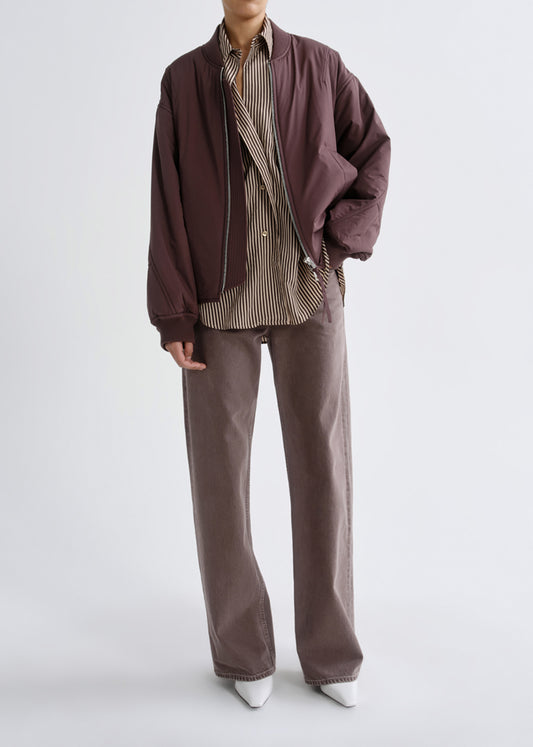 A model is shown from the neck down wearing a dark brown bomber jacket, a brown and beige striped button-down shirt, wide-leg brown pants, and white pointed shoes, against a light gray studio background.