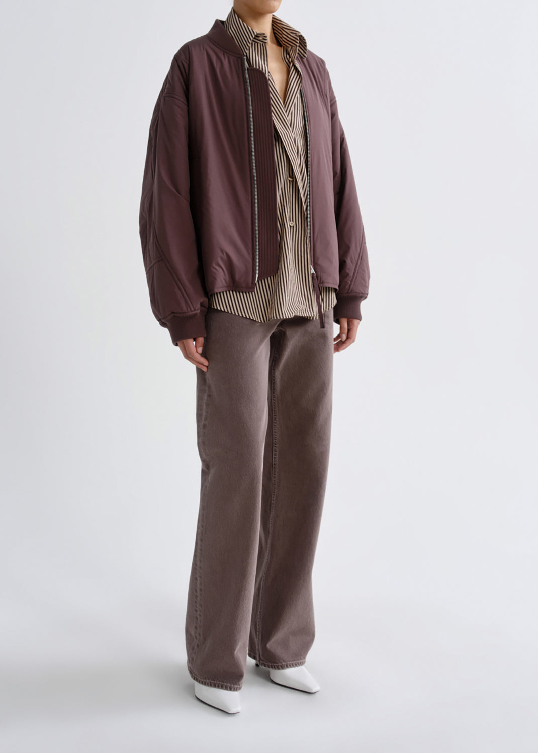 A model is shown from the neck down wearing a dark brown bomber jacket, a beige and brown striped button-down shirt, wide-leg brown pants, and white pointed shoes, against a light gray studio background.