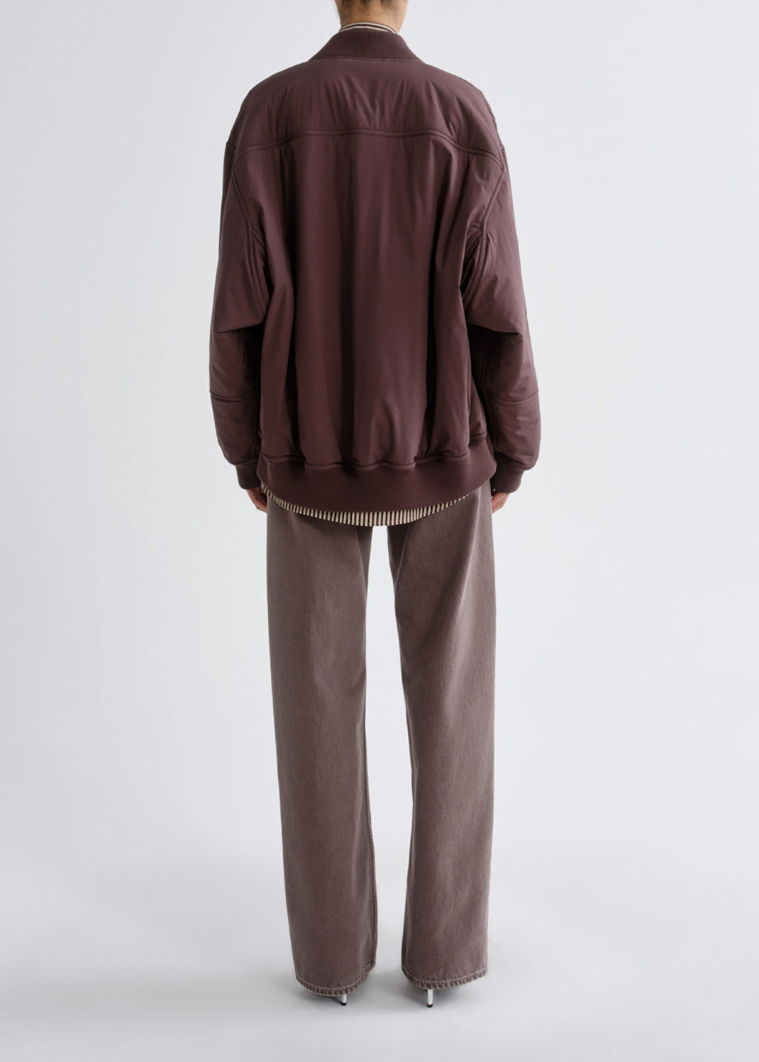 A model shown from the back wearing a dark brown bomber jacket and matching wide-leg trousers against a light gray studio background.