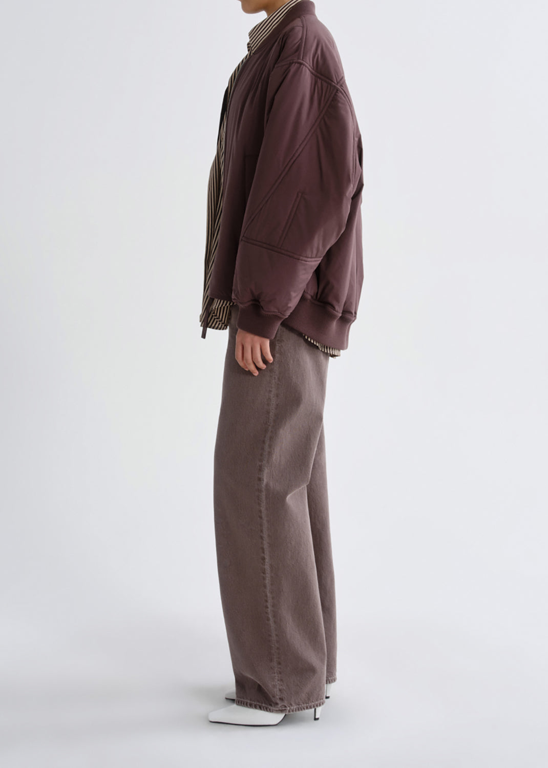 A model shown from the side wearing a dark brown quilted bomber jacket, wide-leg taupe pants, and white pointed-toe heels against a plain light gray studio background.