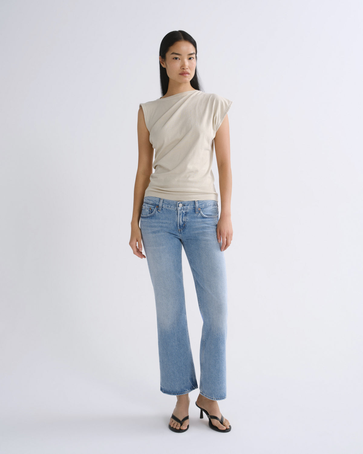A model stands against a light gray studio background wearing a beige asymmetrical sleeveless top, light-wash blue cropped flare jeans, and black heeled sandals.