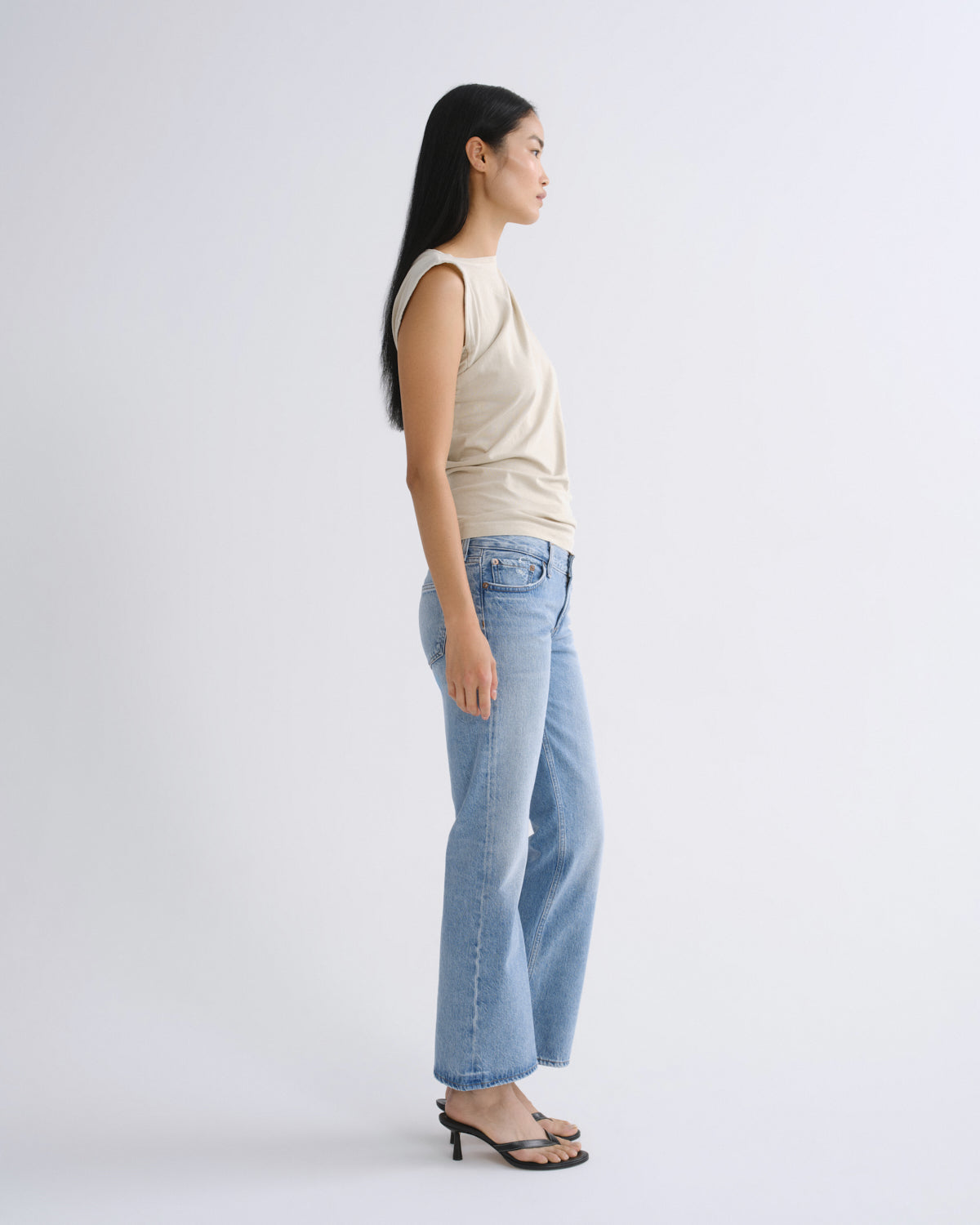 A woman stands in profile wearing a light beige sleeveless top, light blue wide-leg jeans, and black heeled sandals against a plain light gray studio background.