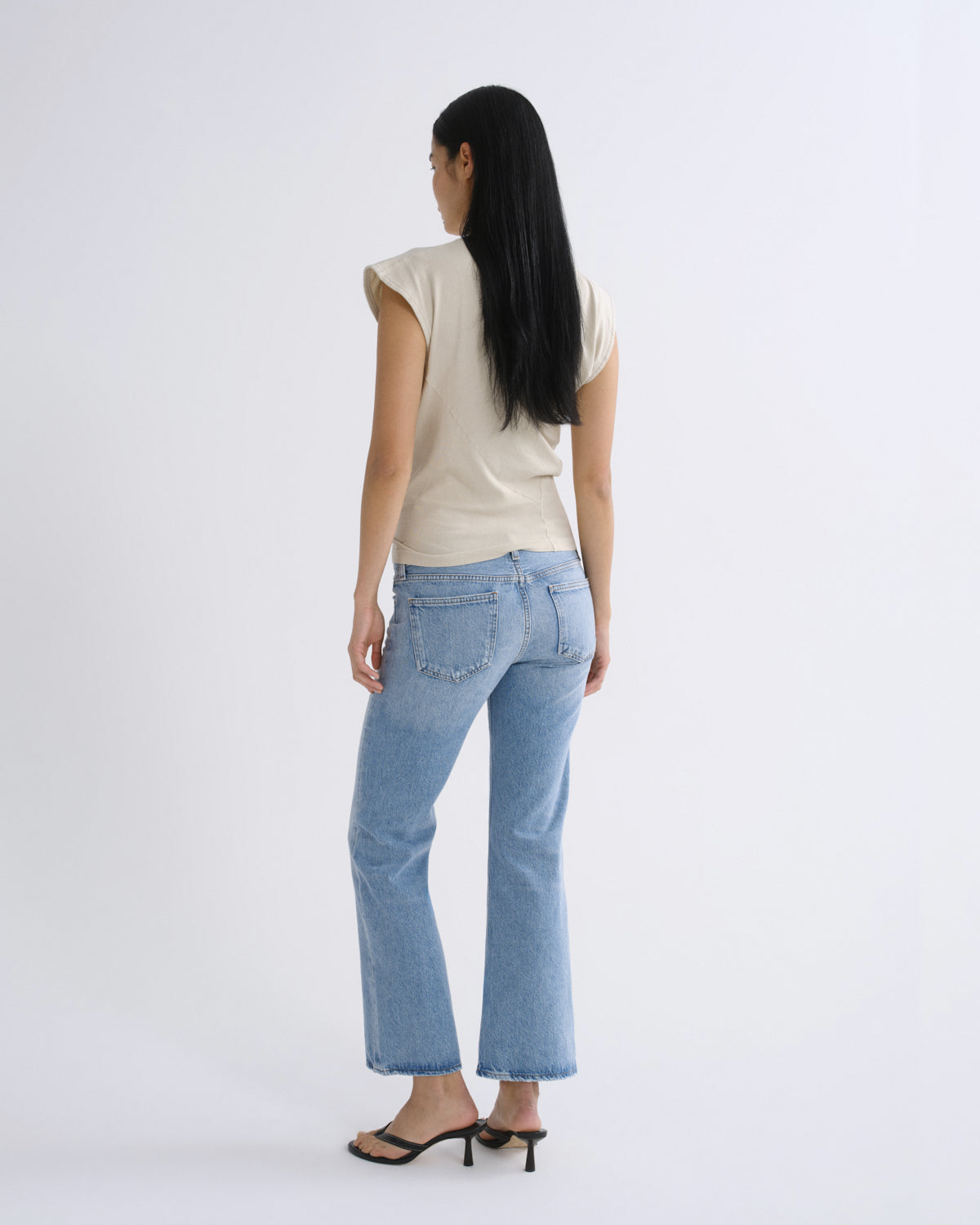 A woman with long black hair is shown from the back wearing light-wash blue cropped flare jeans, a cream short-sleeve top, and black heeled sandals against a plain white studio background.