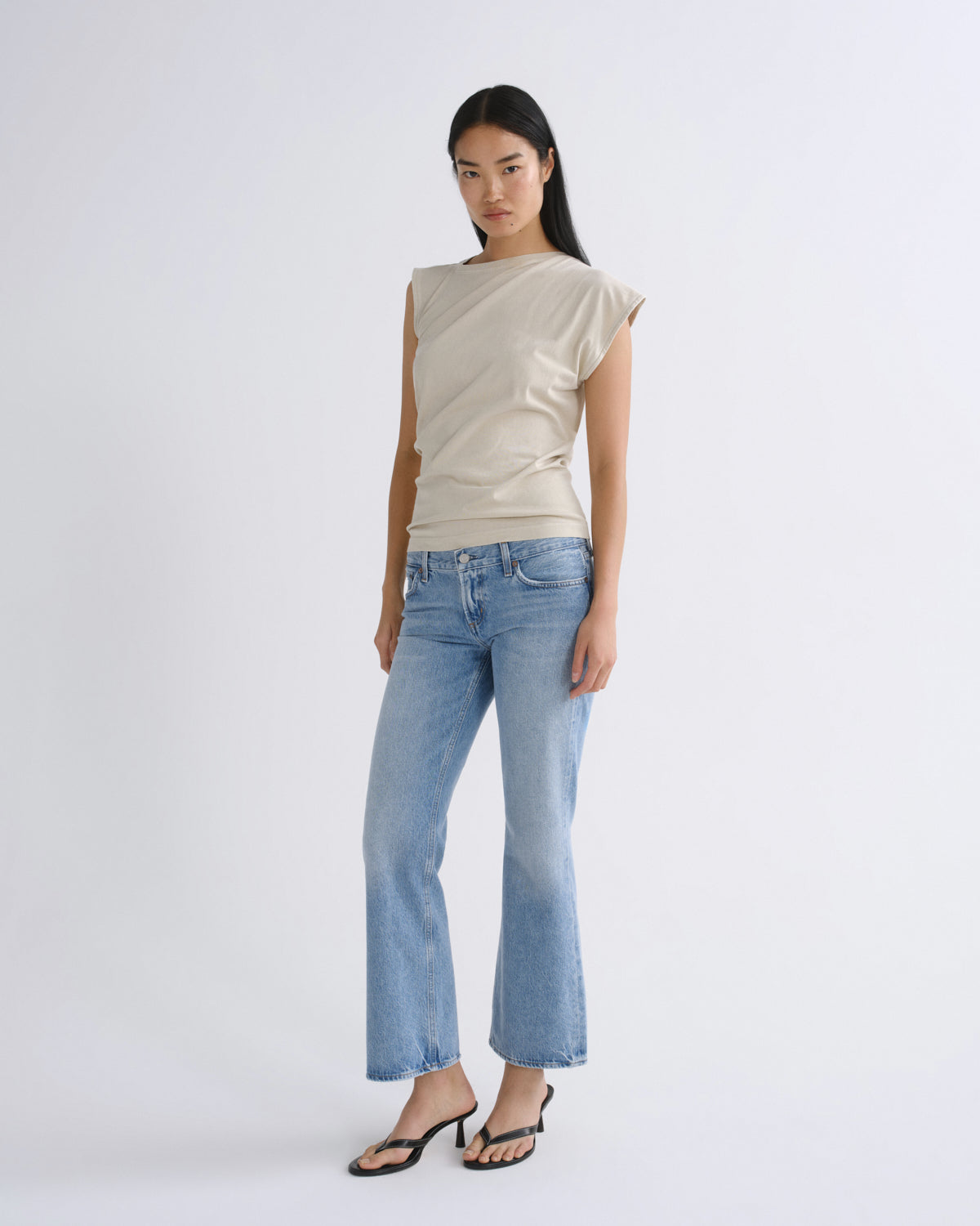 A woman modeling a beige asymmetrical sleeveless top and light-wash blue jeans against a plain light gray studio background.
