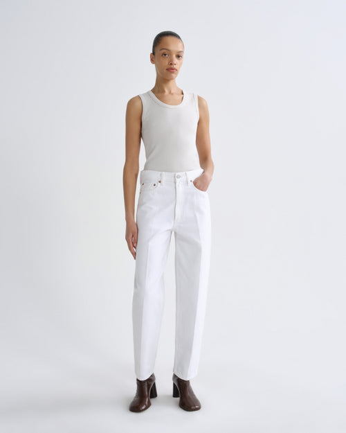 A model stands against a light gray studio background wearing a fitted sleeveless cream top, white high-waisted straight-leg jeans, and dark brown heeled ankle boots.
