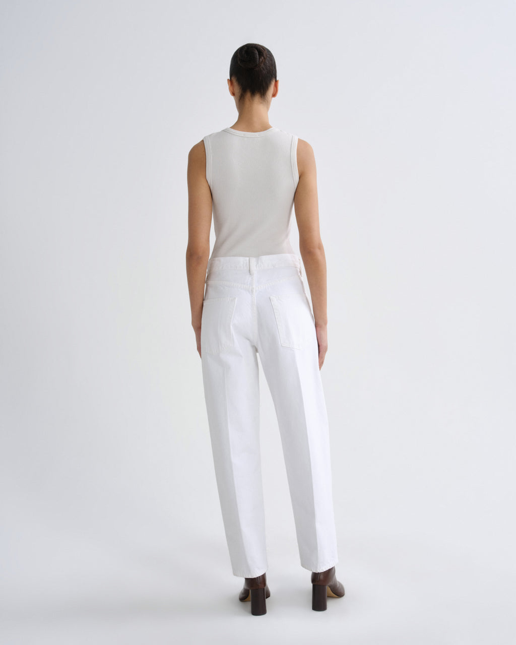 A model viewed from the back wearing a sleeveless light beige top and white trousers, standing against a plain light gray studio background.