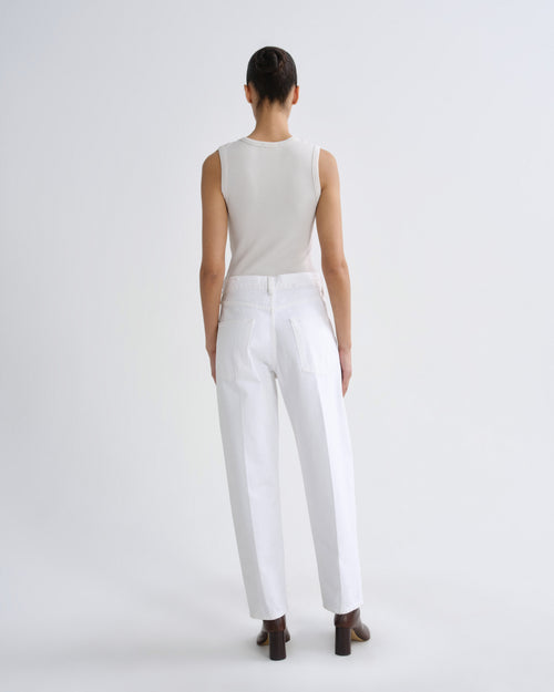 A model viewed from the back wearing a sleeveless light beige top and white trousers, standing against a plain light gray studio background.