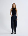 A woman stands against a light gray studio background wearing dark blue high-waisted straight-leg jeans, a black sleeveless top, and black ankle boots.