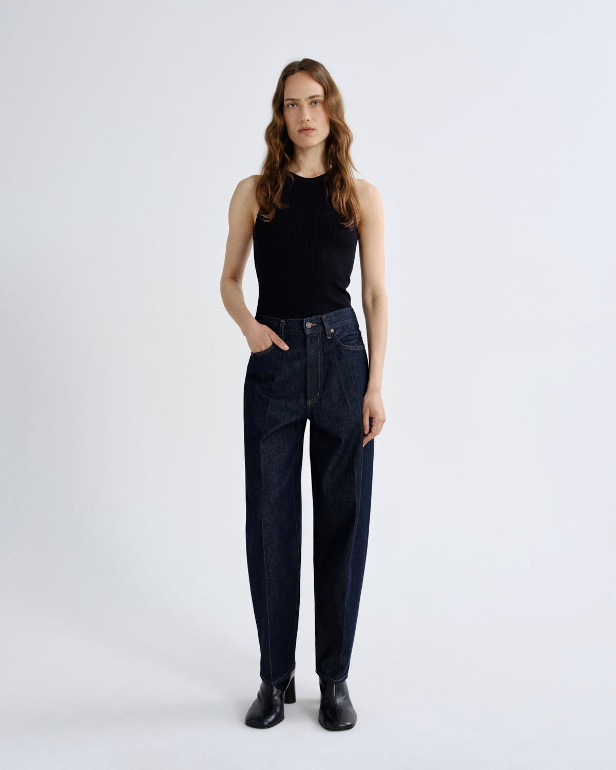 A woman stands against a light gray studio background wearing dark blue high-waisted straight-leg jeans, a black sleeveless top, and black ankle boots.