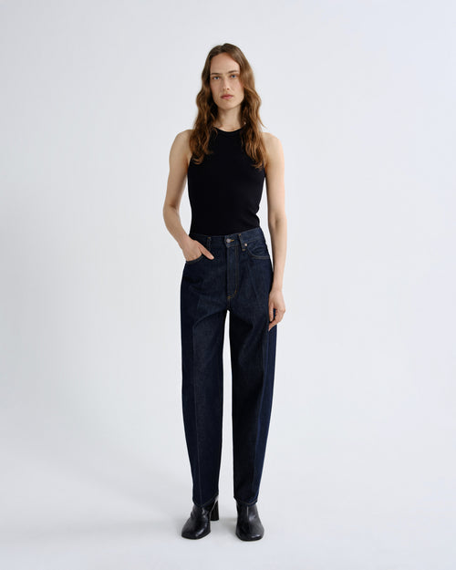 A woman stands against a light gray studio background wearing dark blue high-waisted straight-leg jeans, a black sleeveless top, and black ankle boots.