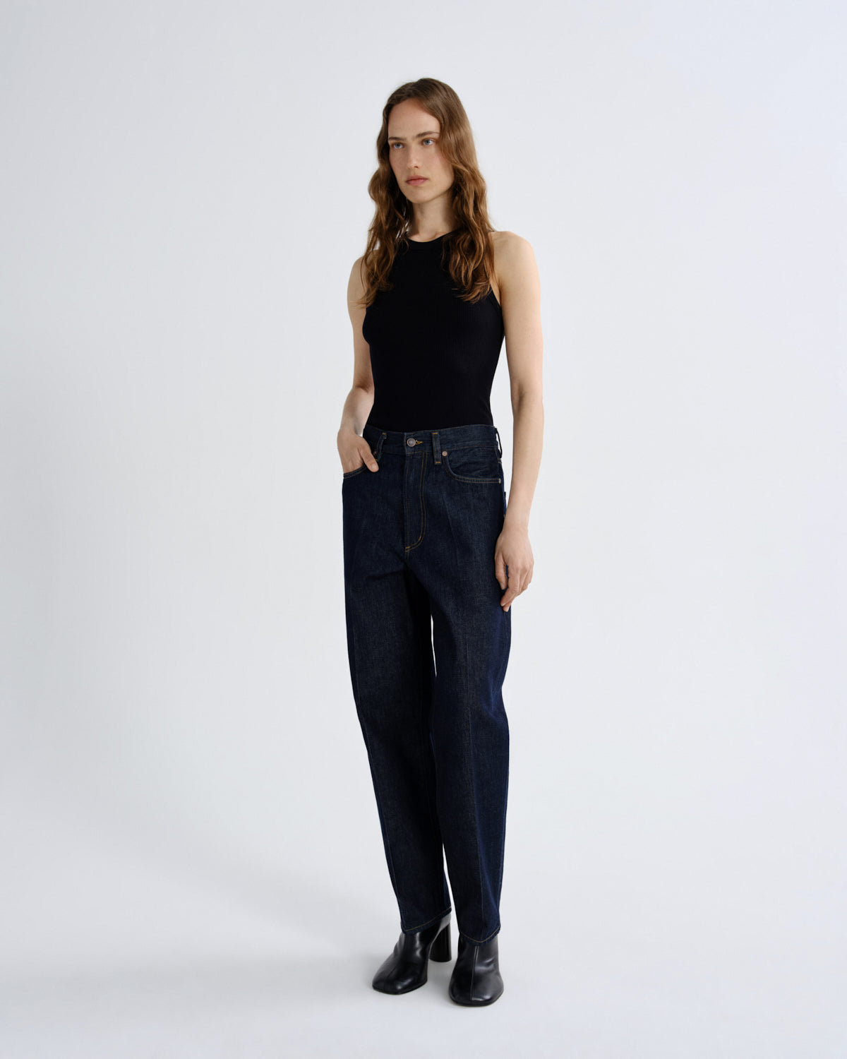 A woman modeling dark wash straight-leg jeans, paired with a black sleeveless top and black boots, standing against a plain light gray studio background.