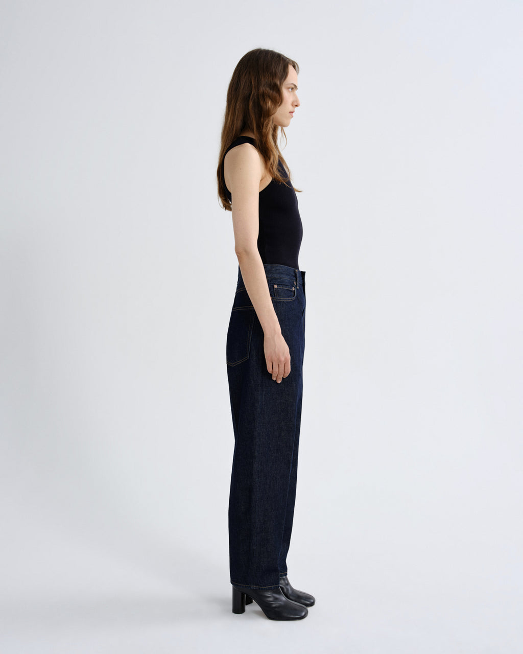 A woman stands in profile wearing a black sleeveless top, dark blue wide-leg jeans, and black heeled shoes against a plain light gray studio background.