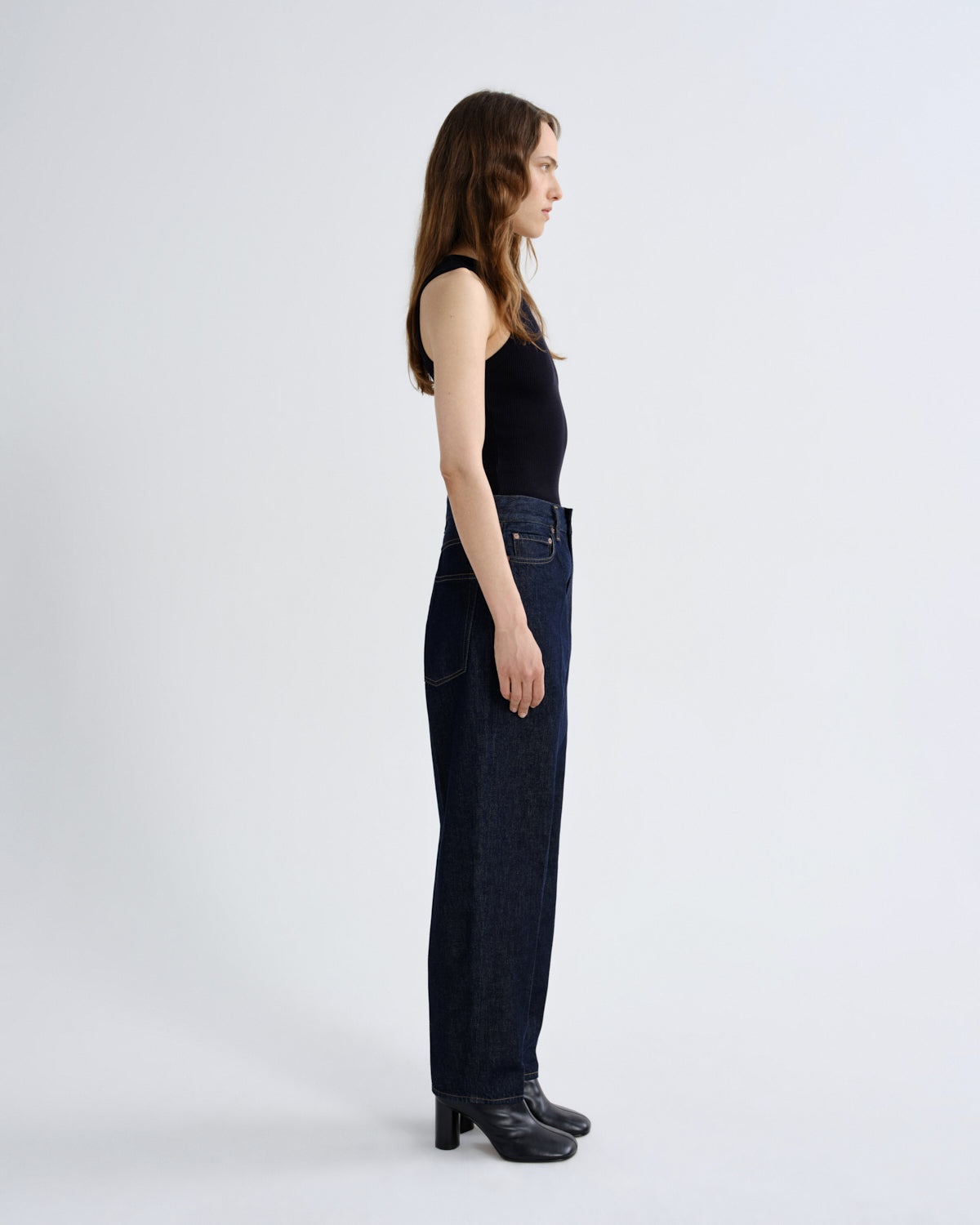A woman stands in profile wearing a black sleeveless top, dark blue wide-leg jeans, and black heeled shoes against a plain light gray studio background.