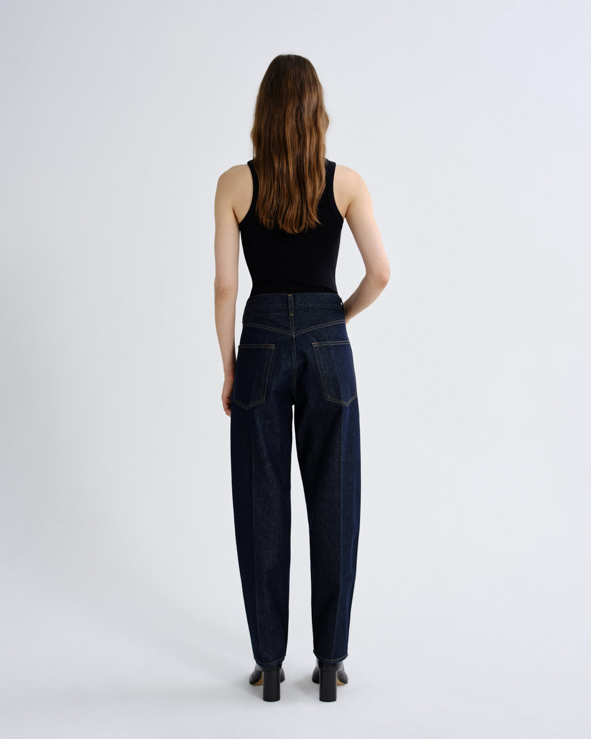 A woman viewed from the back wearing dark blue high-waisted wide-leg jeans, a black tank top, and black heels, standing against a light gray studio background.