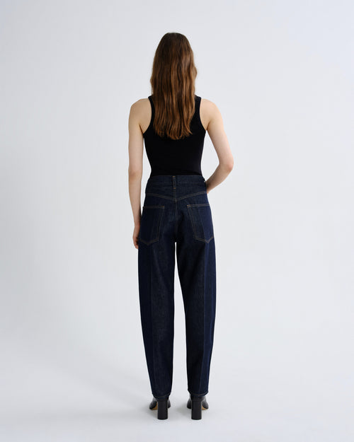 A woman viewed from the back wearing dark blue high-waisted wide-leg jeans, a black tank top, and black heels, standing against a light gray studio background.