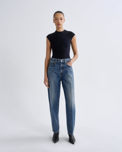 A model stands against a light gray studio background wearing a black sleeveless mock-neck top, blue high-waisted straight-leg jeans, and black pointed ankle boots.