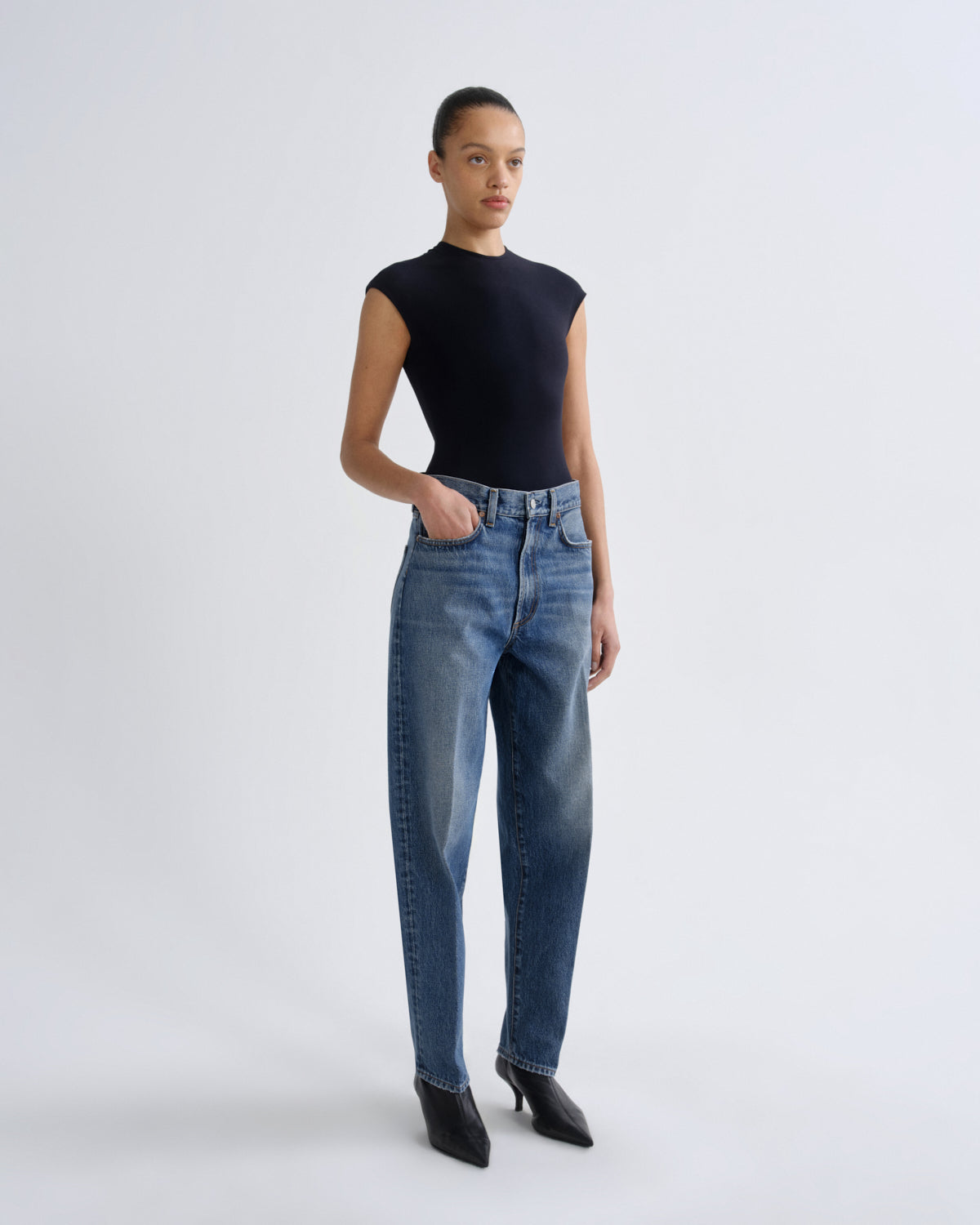 A model stands against a light gray studio background wearing a fitted black sleeveless top, blue high-waisted jeans, and black pointed-toe heels.