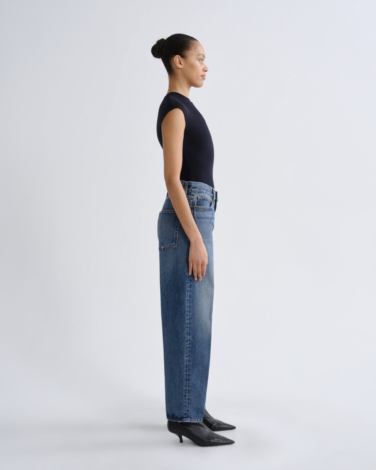 A model in profile wearing a black sleeveless top, wide-leg blue jeans, and black pointed-toe heels, standing against a light gray studio background.