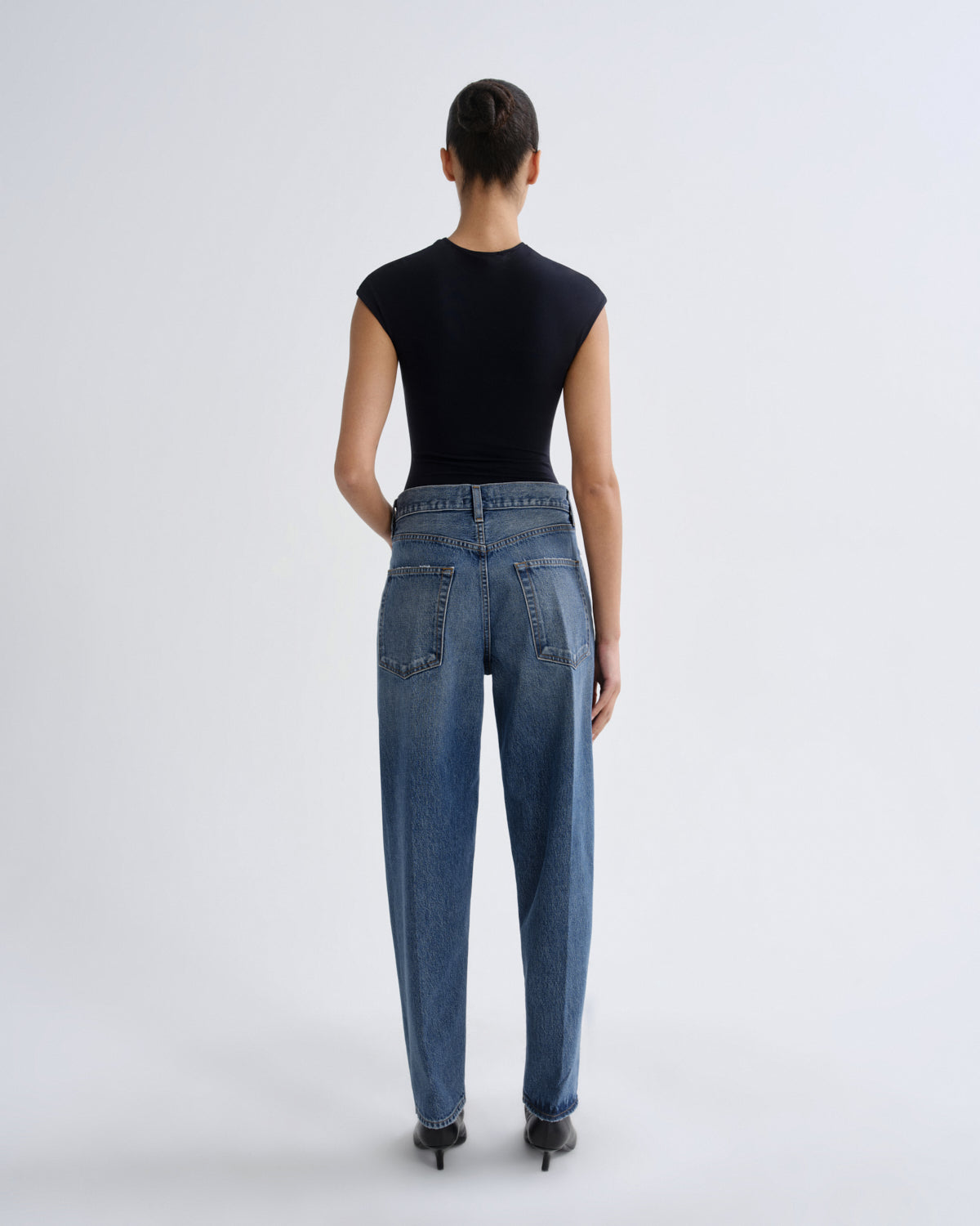 A model viewed from the back wearing a black sleeveless top, blue high-waisted straight-leg jeans, and black pointed-toe heels against a light gray studio background.