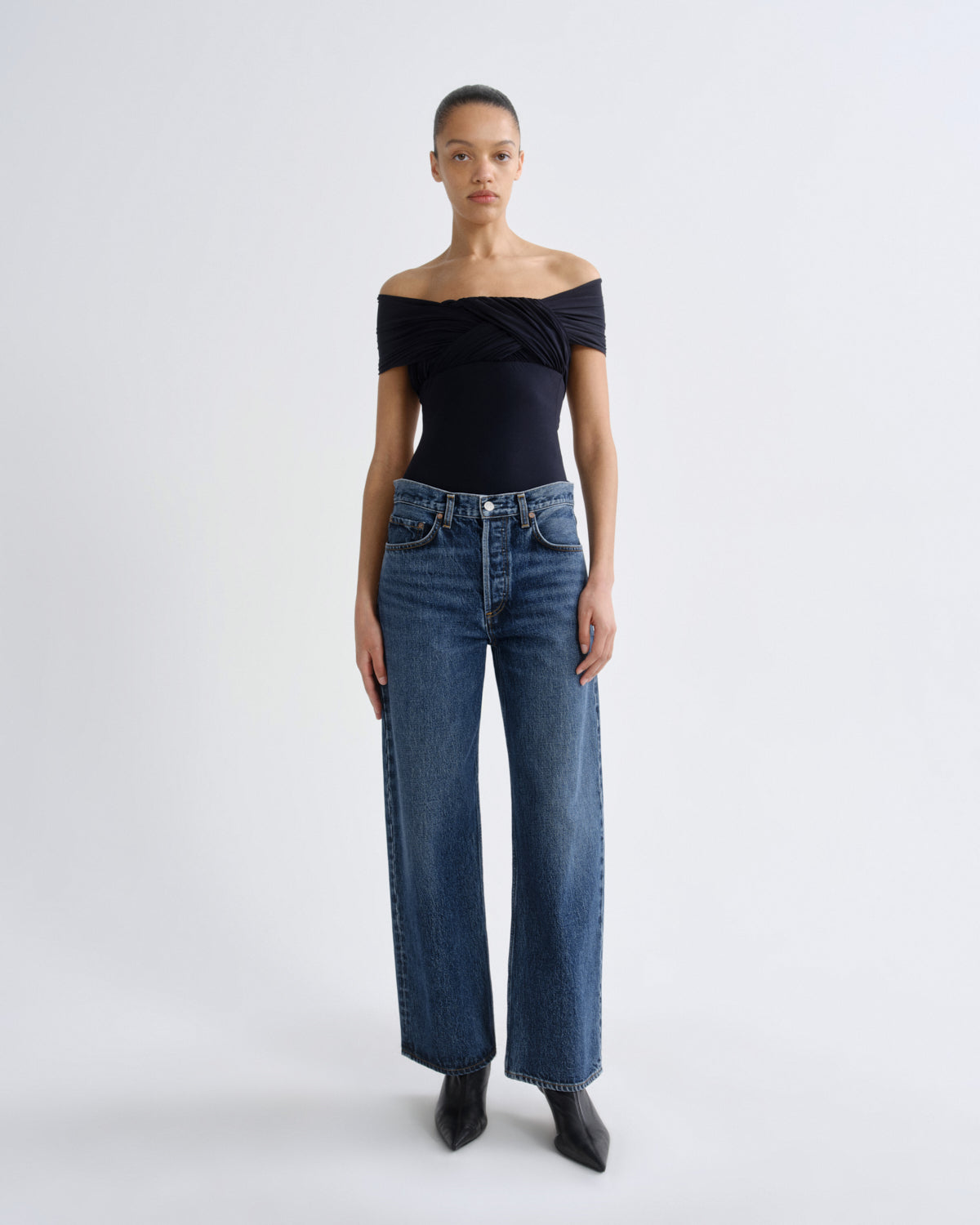 A model stands against a light gray studio background wearing a black off-the-shoulder top, blue wide-leg jeans, and black pointed boots.