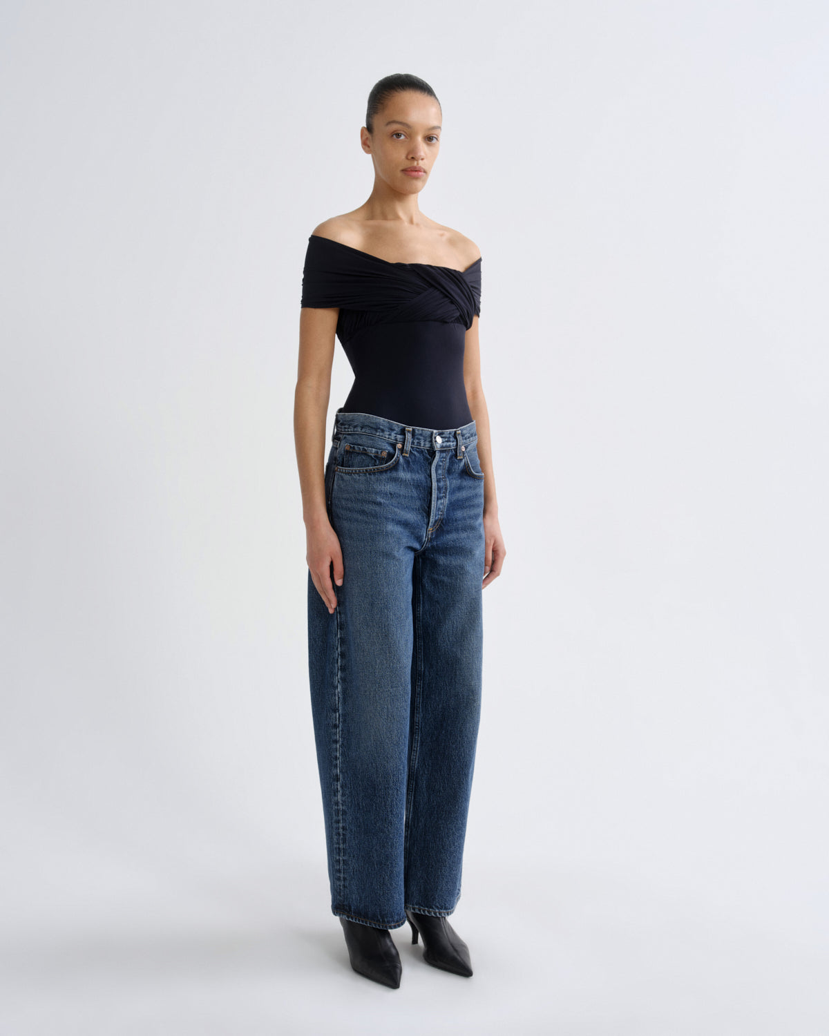 A model stands against a light gray studio background wearing a black off-the-shoulder top, blue wide-leg jeans, and black pointed shoes.