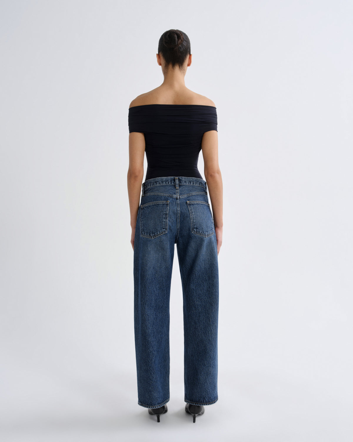 A model shown from the back wearing high-waisted, wide-leg blue jeans and a black off-the-shoulder top against a plain light gray studio background.