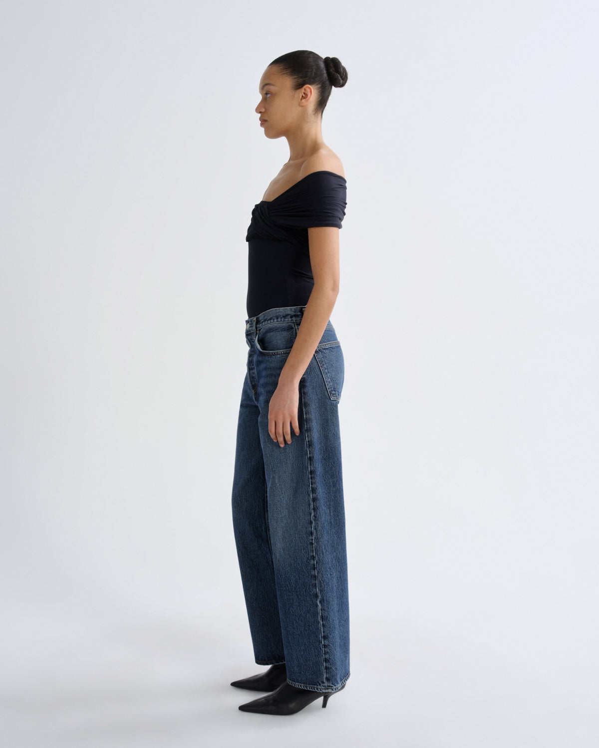A model shown in profile wearing a black off-the-shoulder short-sleeve top, high-waisted wide-leg blue jeans, and black pointed-toe heels, standing against a plain light gray studio background.