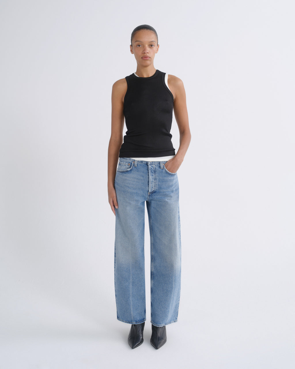 A model stands against a light gray studio background wearing a black sleeveless top with white shoulder panels, light-wash wide-leg blue jeans, and black pointed boots.