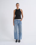 A model stands against a light gray studio background wearing a black sleeveless top with white shoulder panels, light-wash wide-leg blue jeans, and black pointed boots.