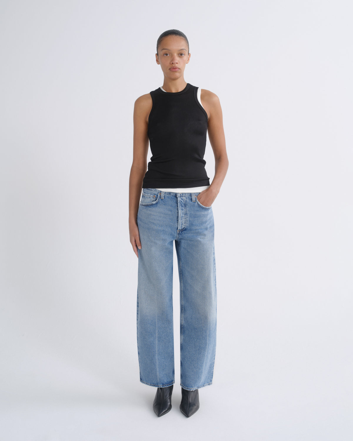 A model stands against a light gray studio background wearing a black sleeveless top with white shoulder panels, light-wash wide-leg blue jeans, and black pointed boots.