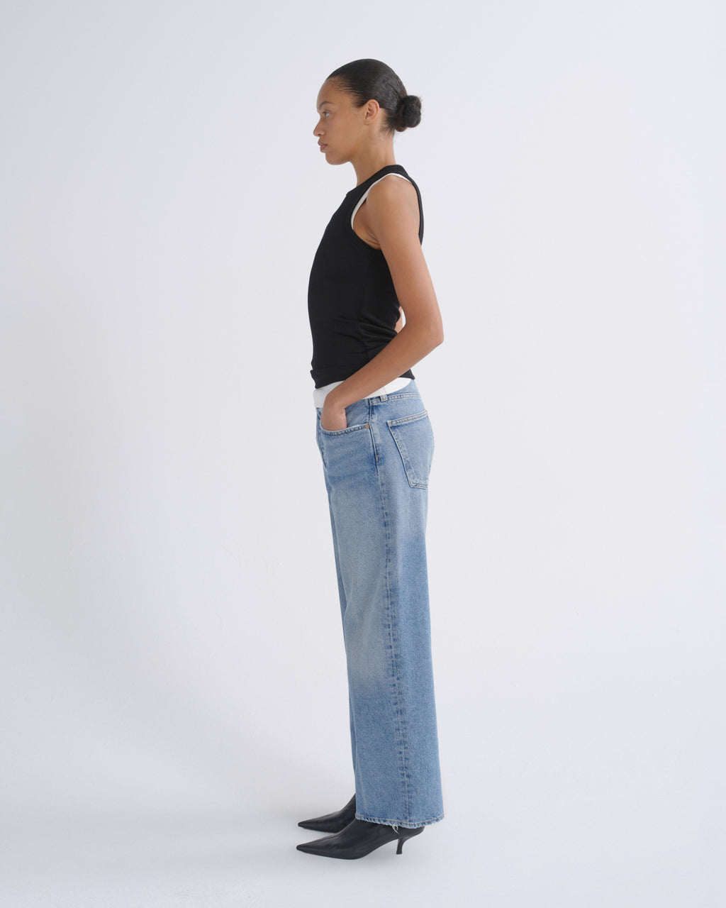 A model in profile wearing a black sleeveless top, wide-leg light blue denim jeans, and black pointed-toe heels, standing against a plain light gray studio background.