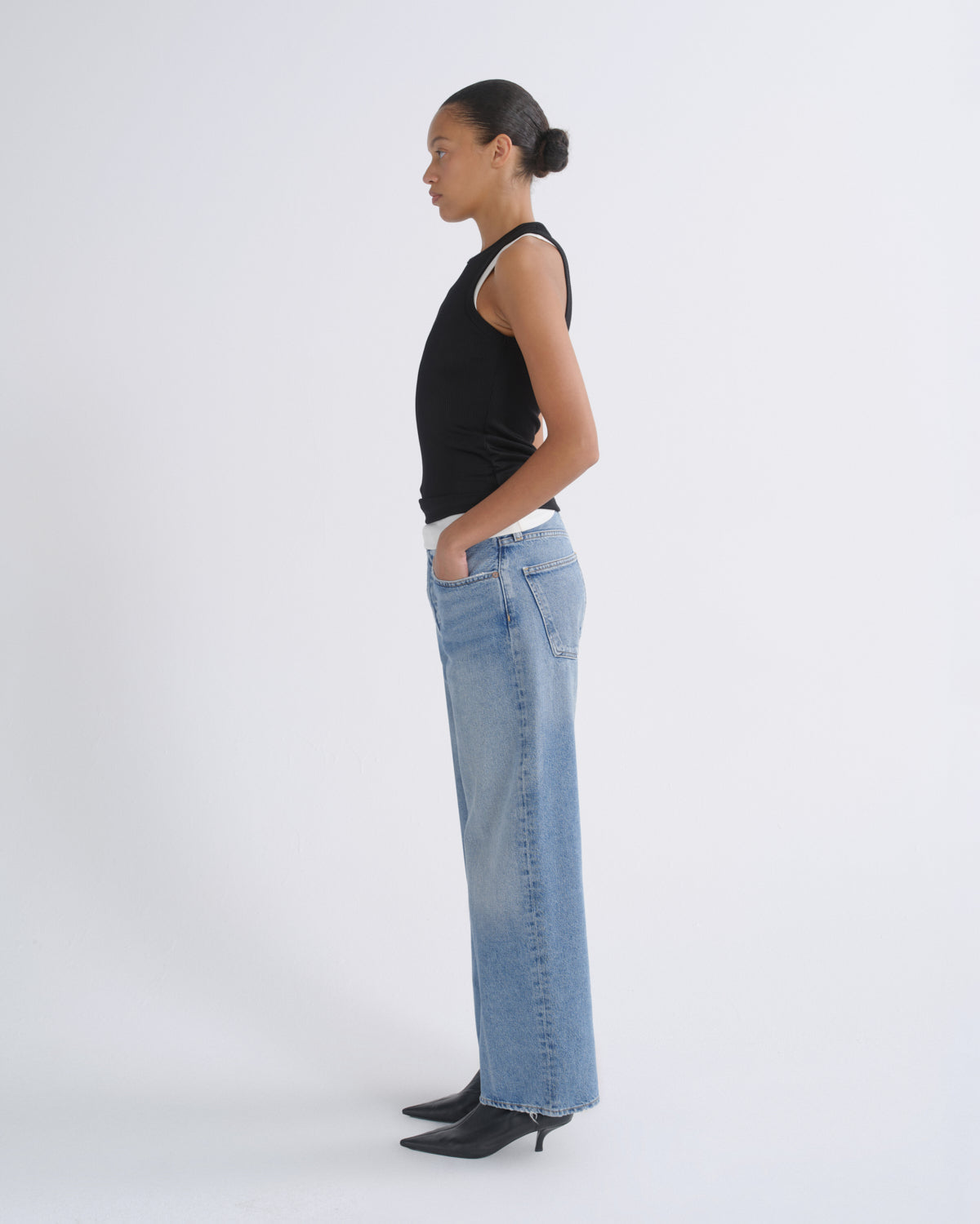 A model in profile wearing a black sleeveless top, wide-leg light blue denim jeans, and black pointed-toe heels, standing against a plain light gray studio background.