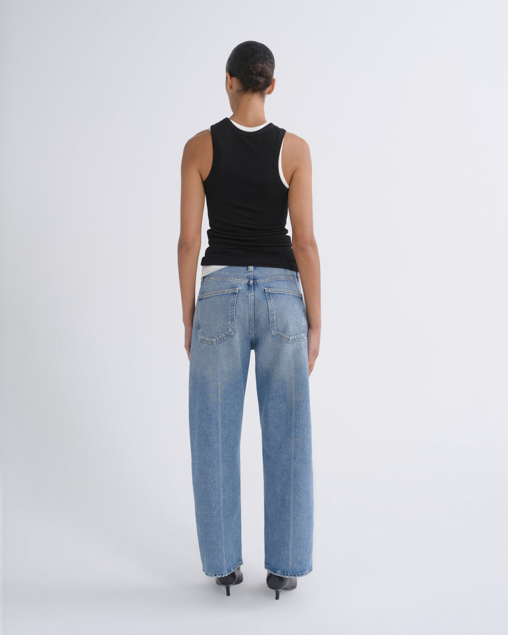 A model viewed from the back wearing a black sleeveless top with a white inner collar and light-wash wide-leg blue jeans, standing against a plain light gray studio background.