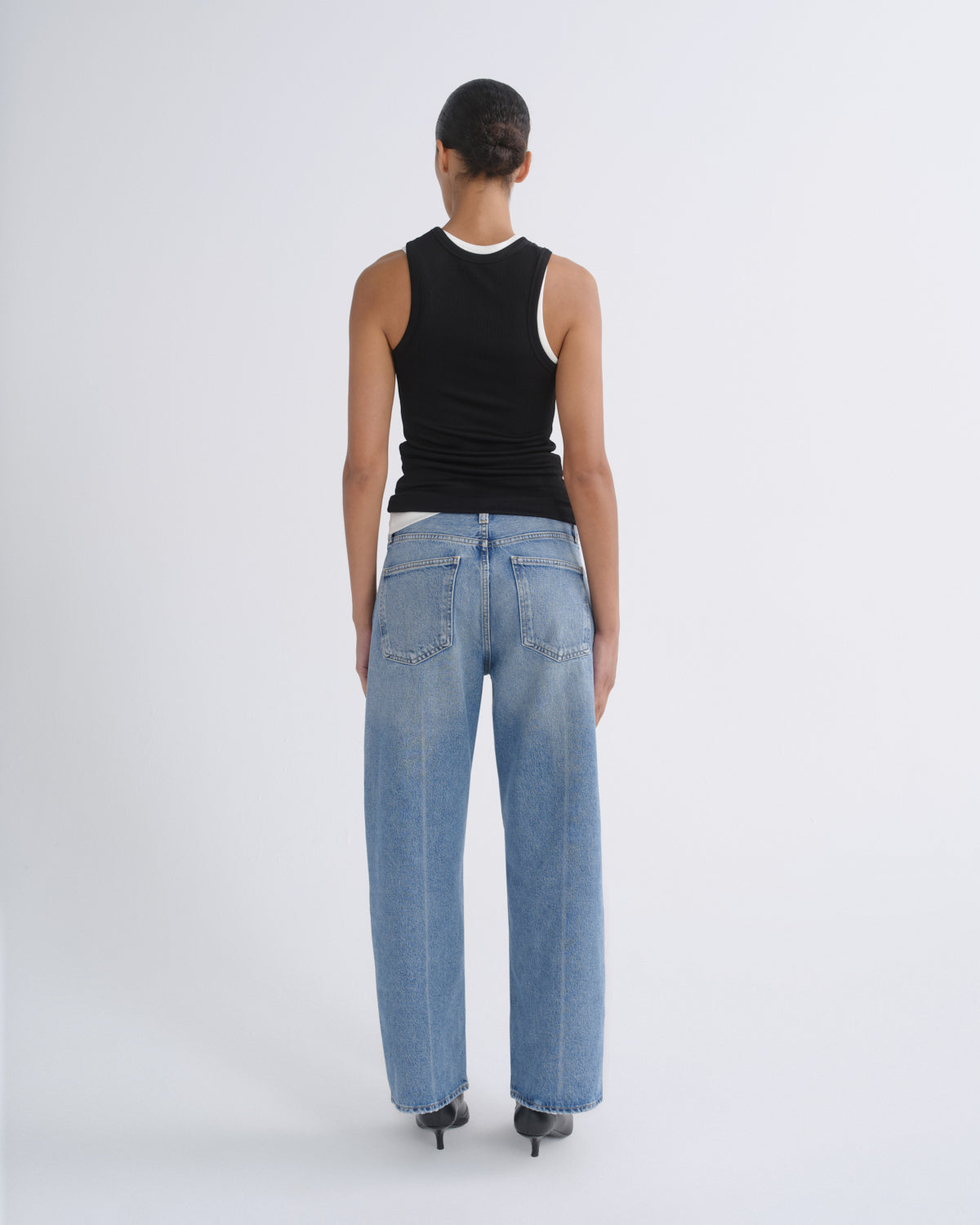 A model viewed from the back wearing a black sleeveless top with a white inner collar and light-wash wide-leg blue jeans, standing against a plain light gray studio background.