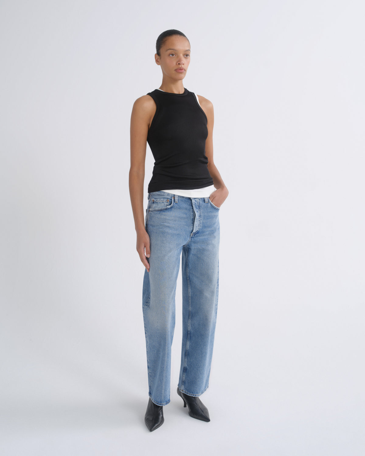 A model stands against a light gray studio background wearing a black sleeveless ribbed top, light-wash blue straight-leg jeans, and black pointed shoes.