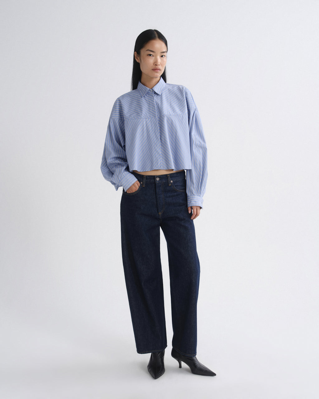 A model stands against a light gray studio background wearing a light blue cropped button-up shirt with a subtle geometric pattern, dark wide-leg jeans, and black pointed-toe heels.