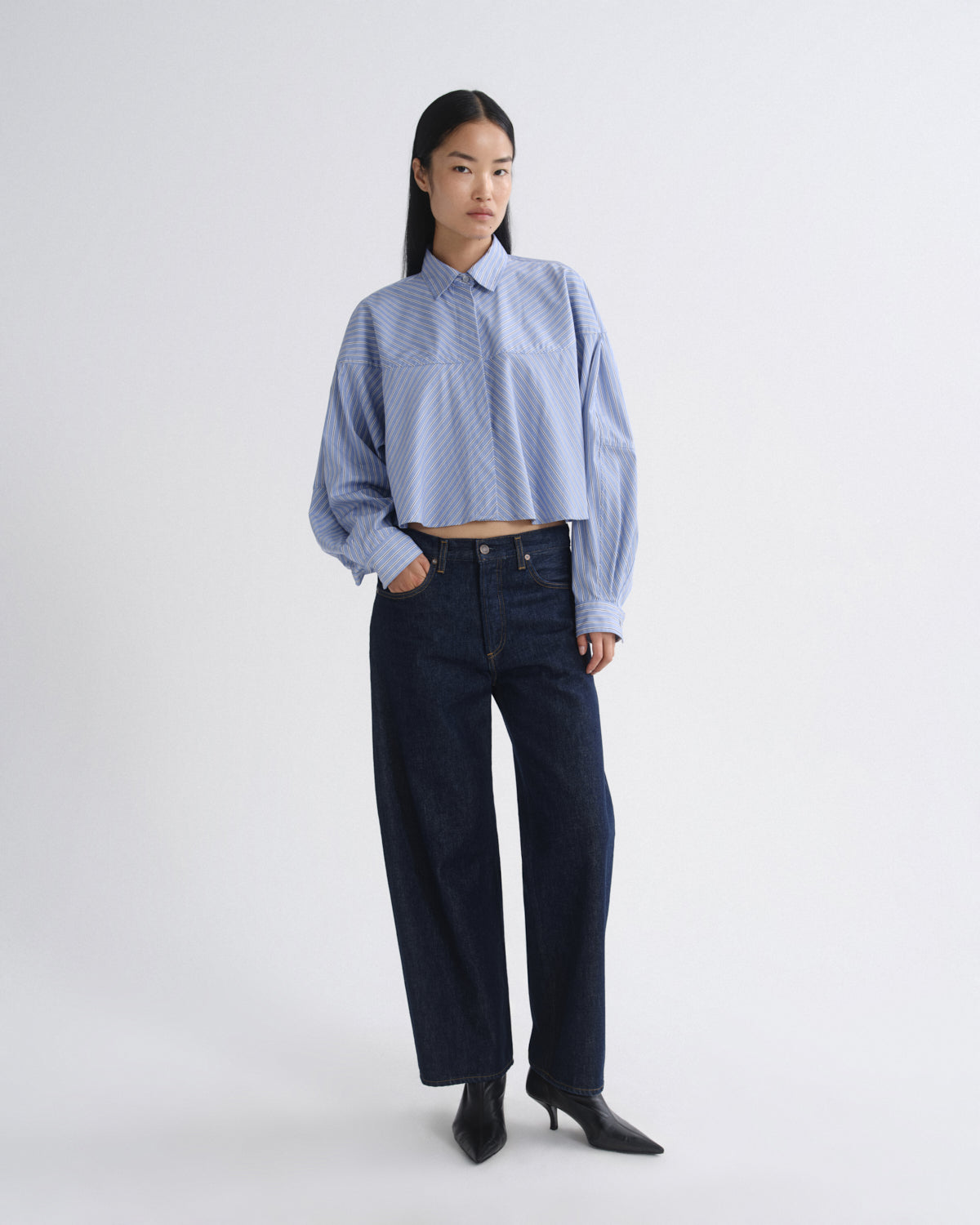 A model stands against a light gray studio background wearing a light blue cropped button-up shirt with a subtle geometric pattern, dark wide-leg jeans, and black pointed-toe heels.