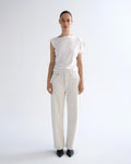A model stands against a plain white studio background wearing a white sleeveless top with ruffled shoulders and white wide-leg jeans, paired with black pointed shoes.