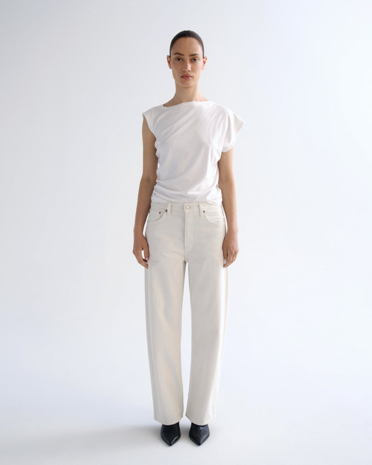 A model stands against a plain white studio background wearing a white sleeveless top with ruffled shoulders and white wide-leg jeans, paired with black pointed shoes.
