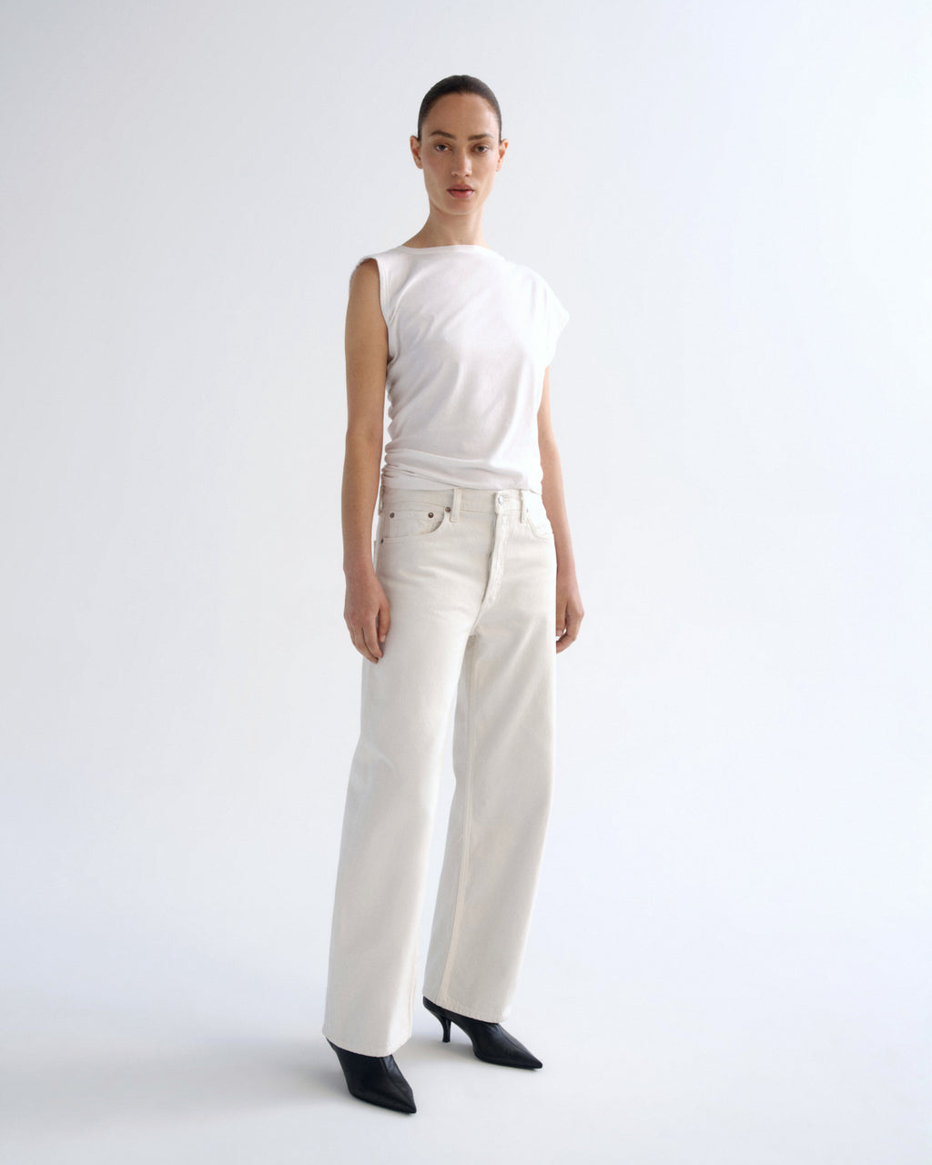 A model stands in a studio wearing a white sleeveless top, off-white wide-leg trousers, and black pointed heels against a plain light background.