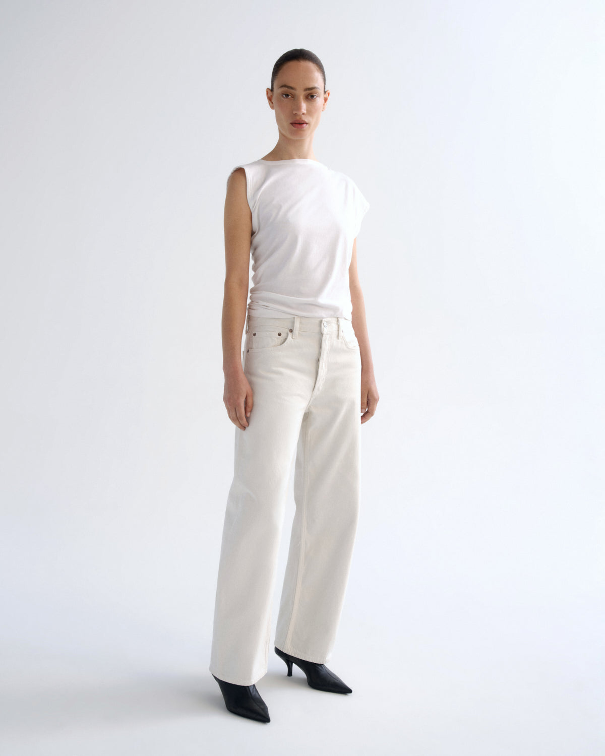 A model stands in a studio wearing a white sleeveless top, off-white wide-leg trousers, and black pointed heels against a plain light background.