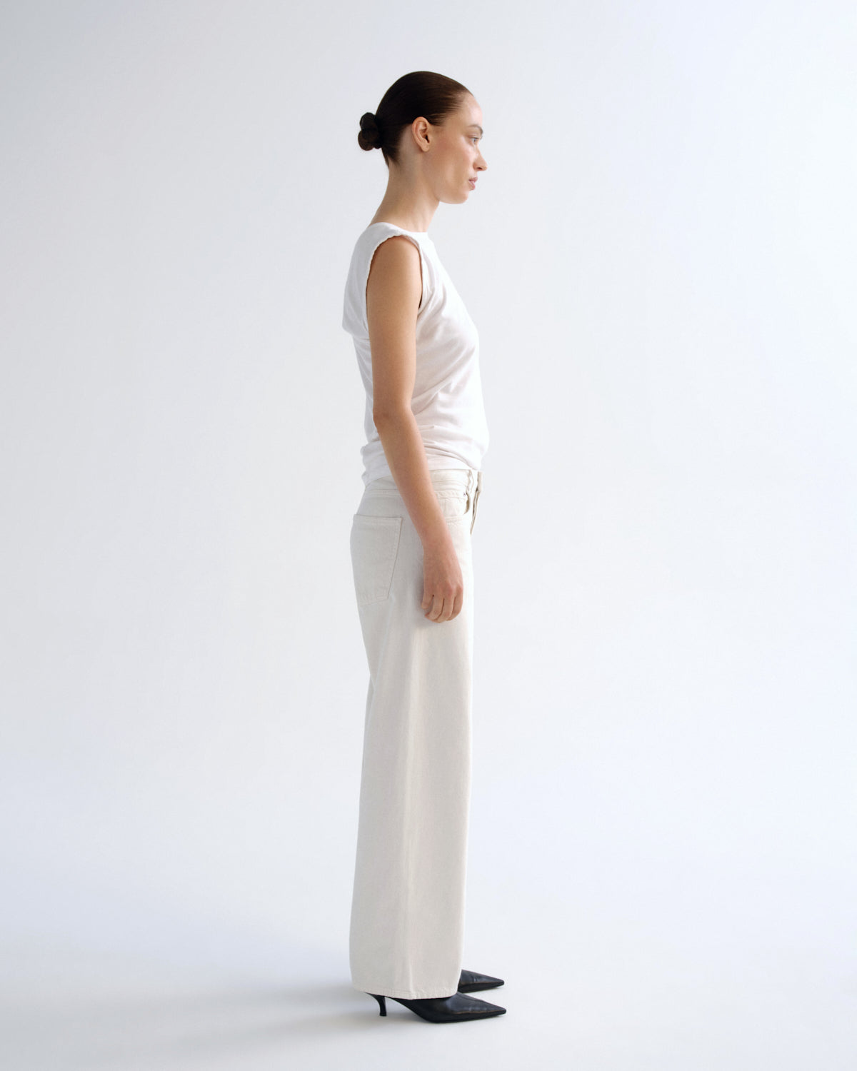 A woman stands in profile wearing a white sleeveless top and white wide-leg trousers against a plain light gray studio background.