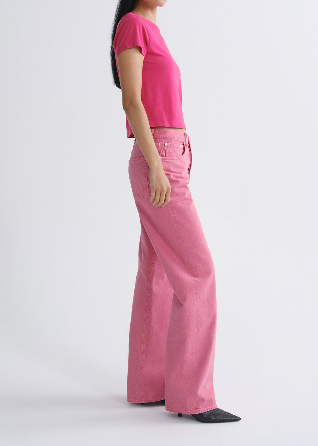 A side-profile view of a model wearing a bright pink short-sleeve crop top and matching wide-leg pink pants against a grey background.